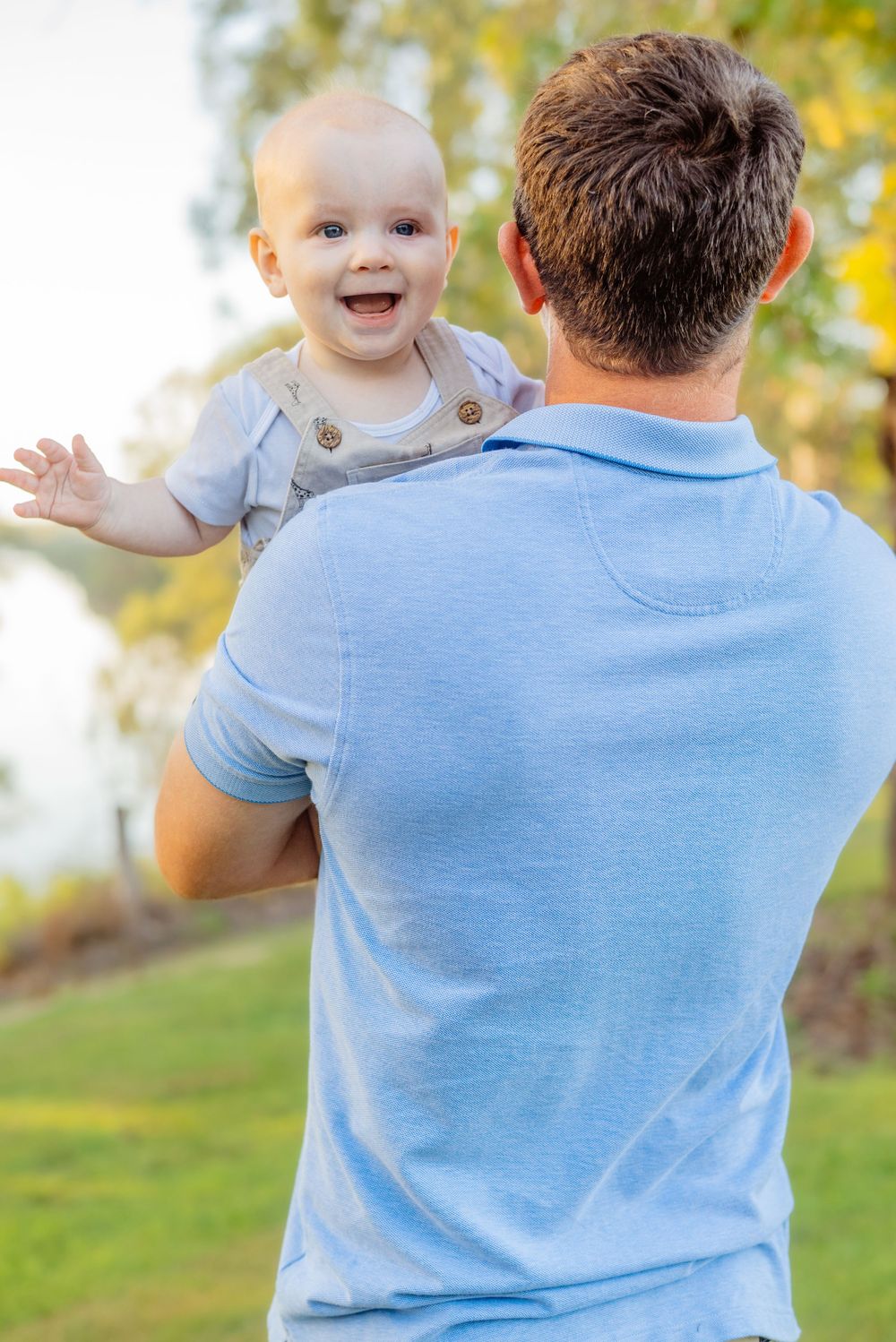 A joyful baby interacts with someone wearing a blue shirt in a sunny park setting.