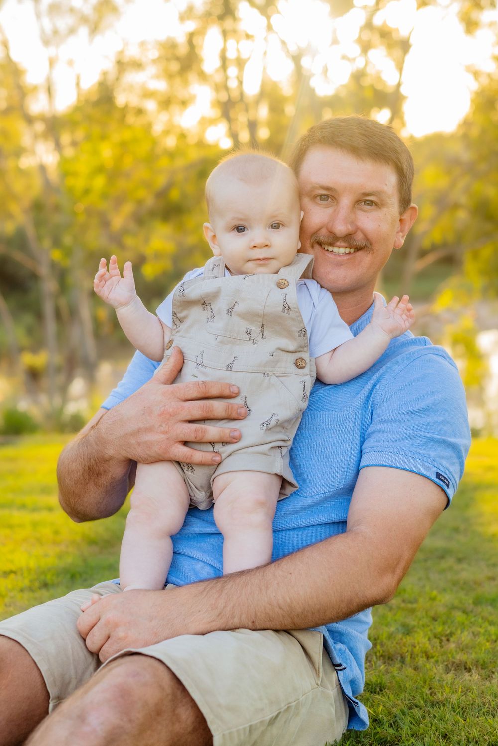 A baby in beige clothing shares happy moments with someone in a blue shirt outdoors.