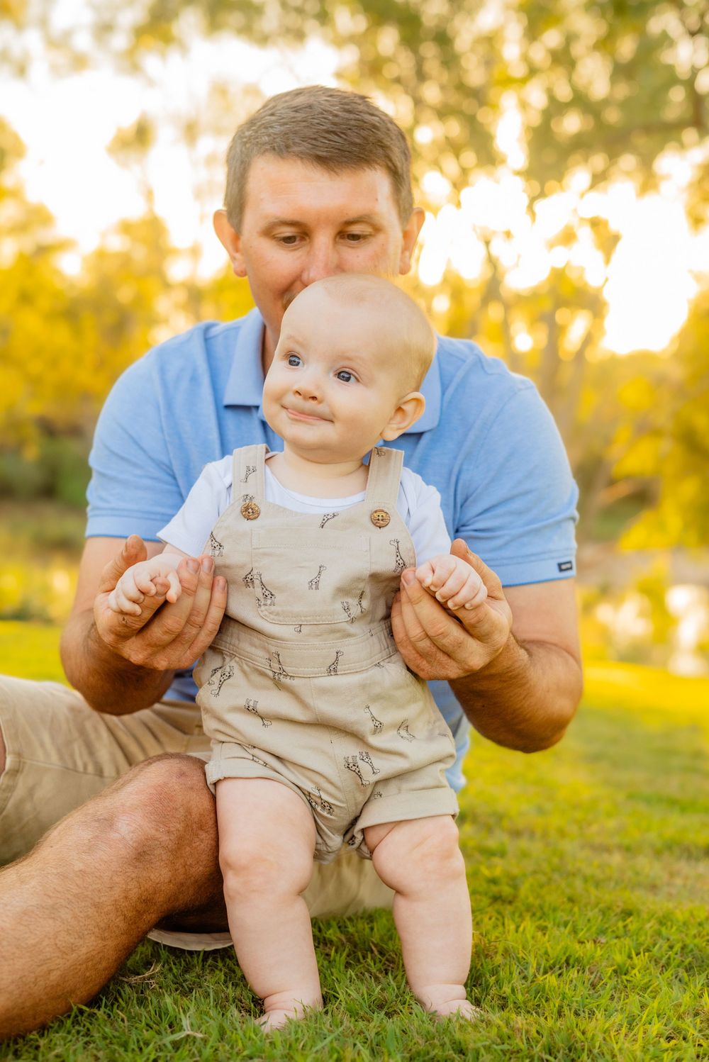 A baby in tan overalls sits on grass with support during golden hour sunset.