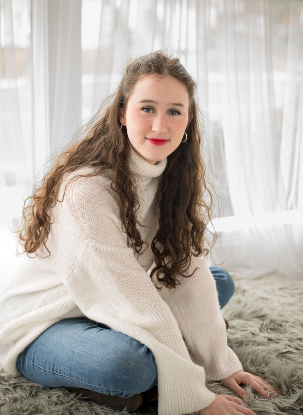 A cozy portrait featuring a cream turtleneck sweater and jeans against white curtains.