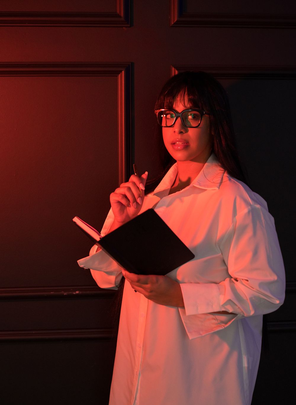 Person in white lab coat holding a clipboard in a dimly lit red room.