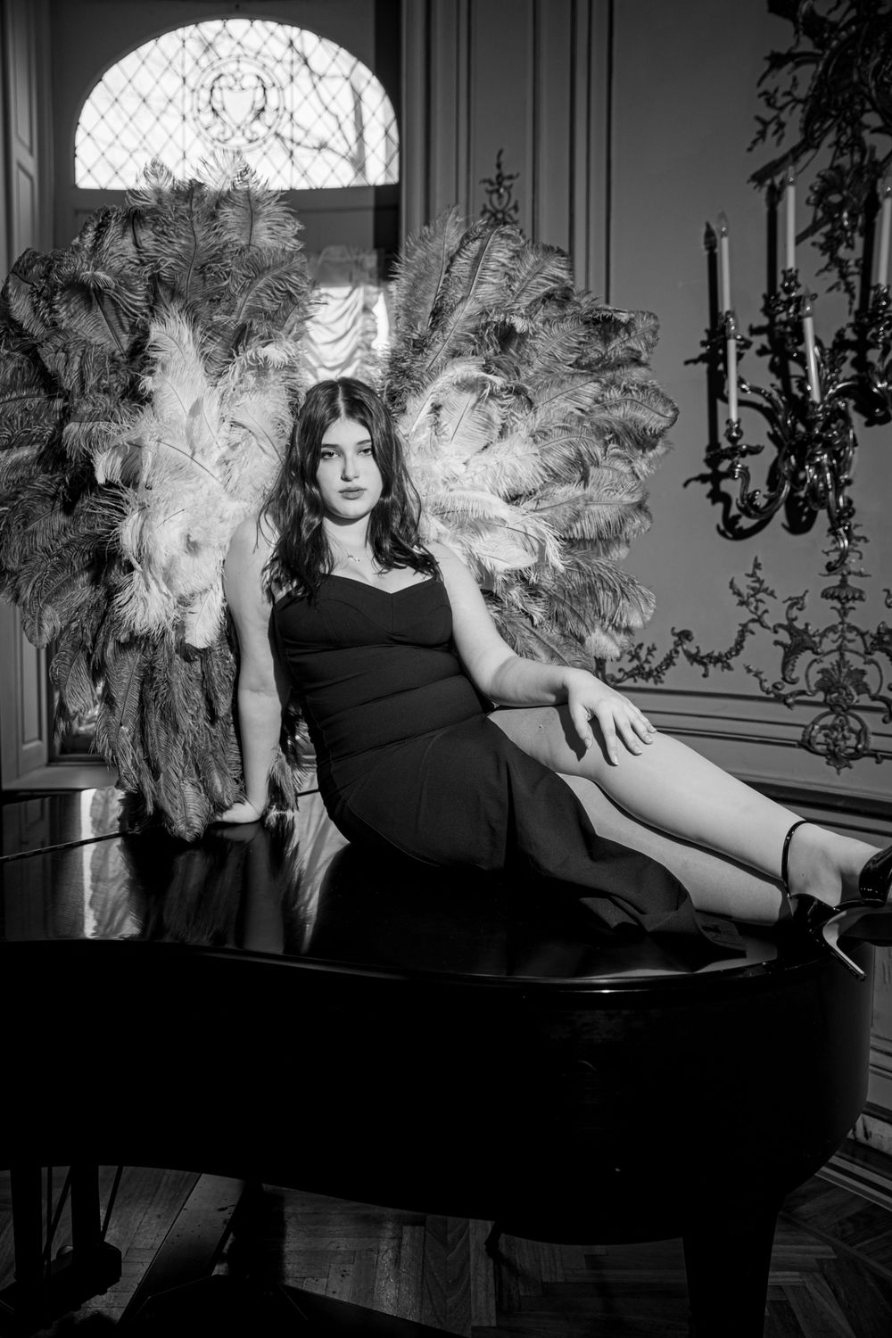 Black and white glamour photo of woman in evening dress reclining on grand piano near ornate window and candelabra.