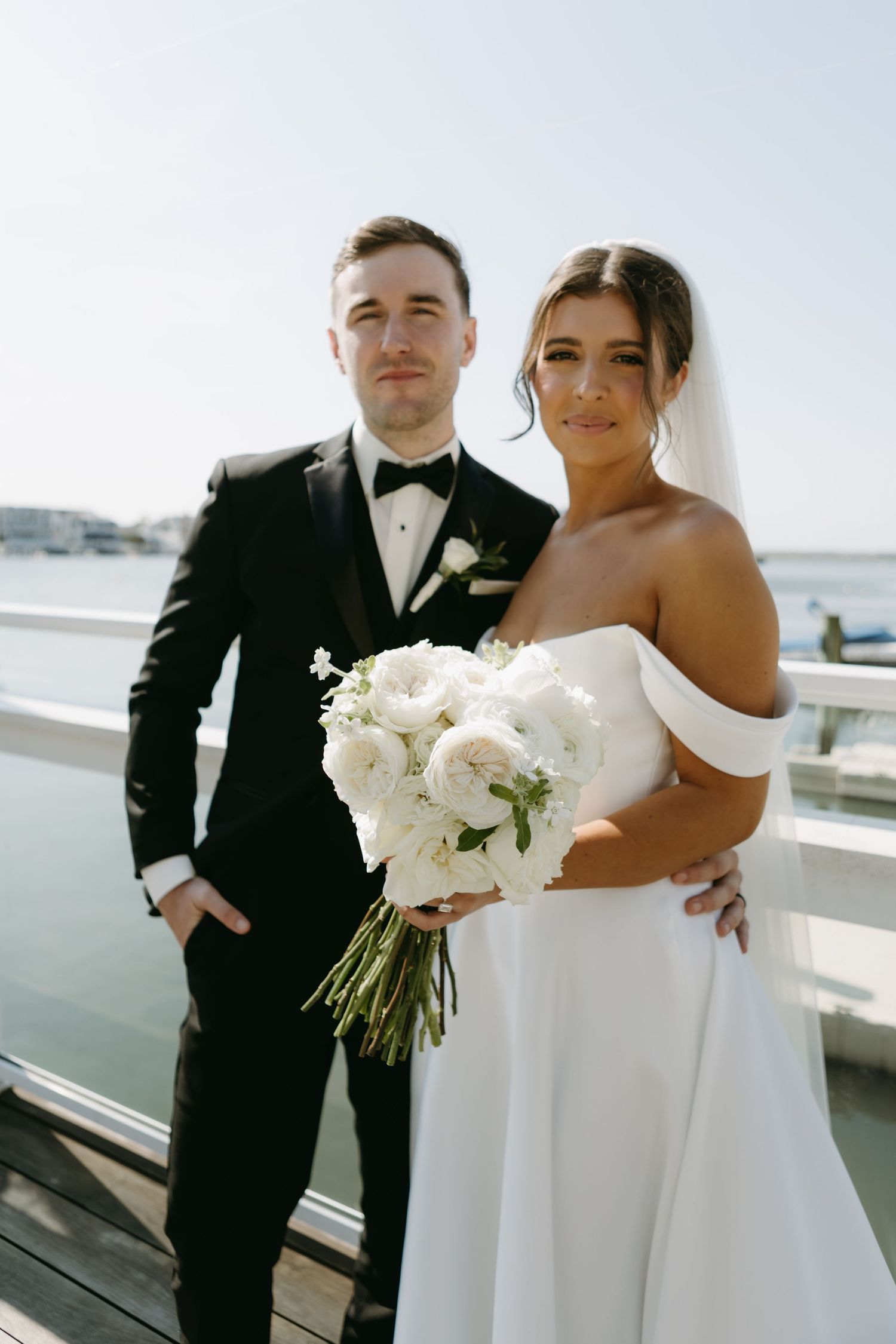 Elegant couple in wedding attire pose together on a waterfront balcony.