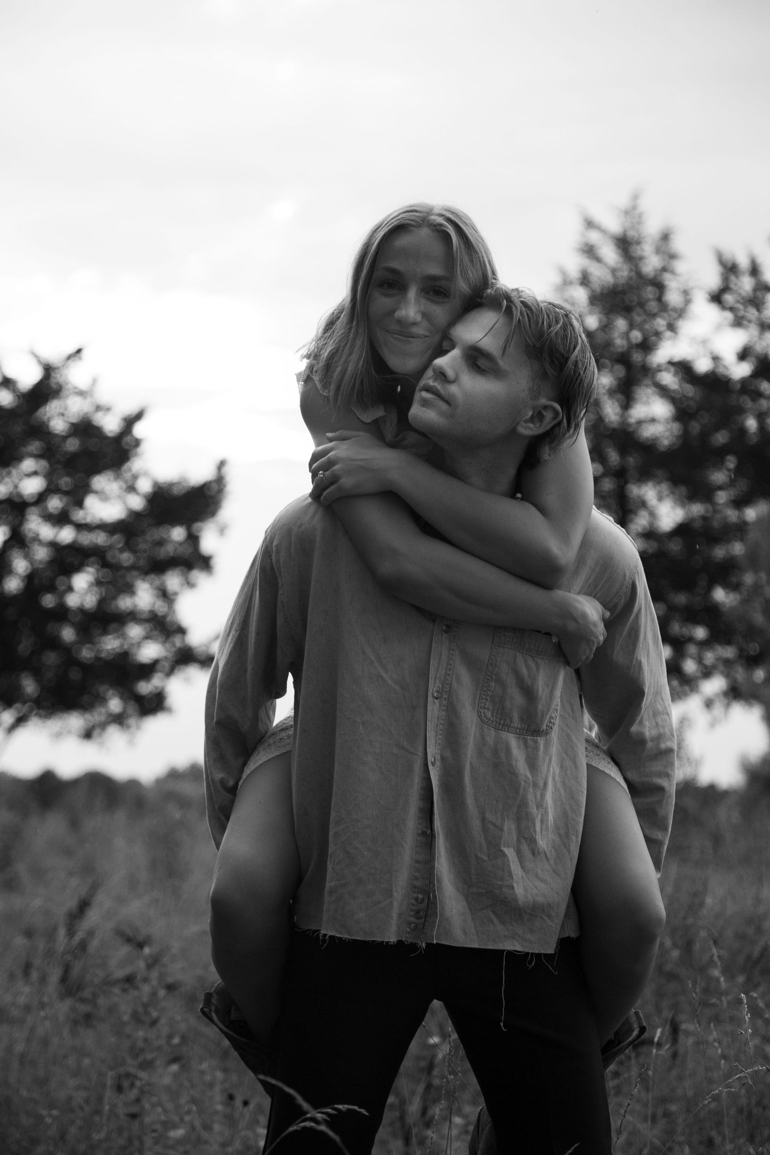 Black and white photo of a couple sharing a tender piggyback moment outdoors with trees in the background at Frank Lisk Park in Charlotte.