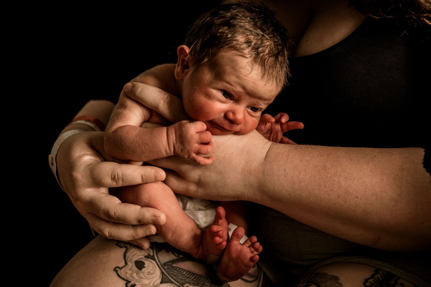 A newborn baby is being held by gentle hands moments after birth in a dark, dramatic setting.
