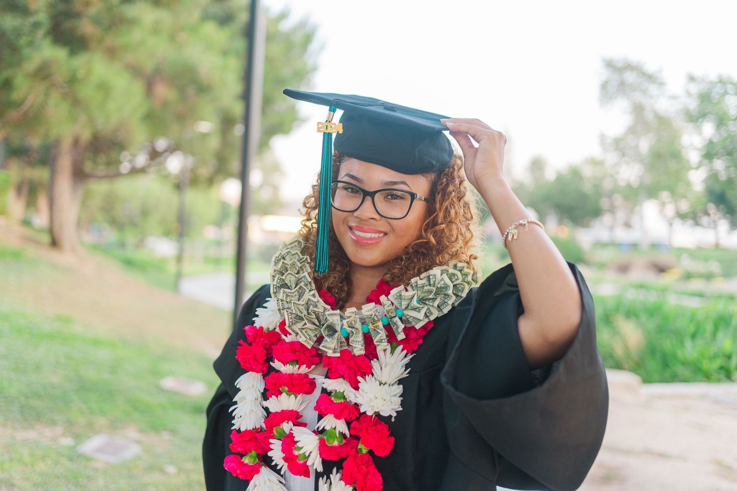 Graduate in cap and gown with multiple colorful flower leis poses outdoors at graduation ceremony.