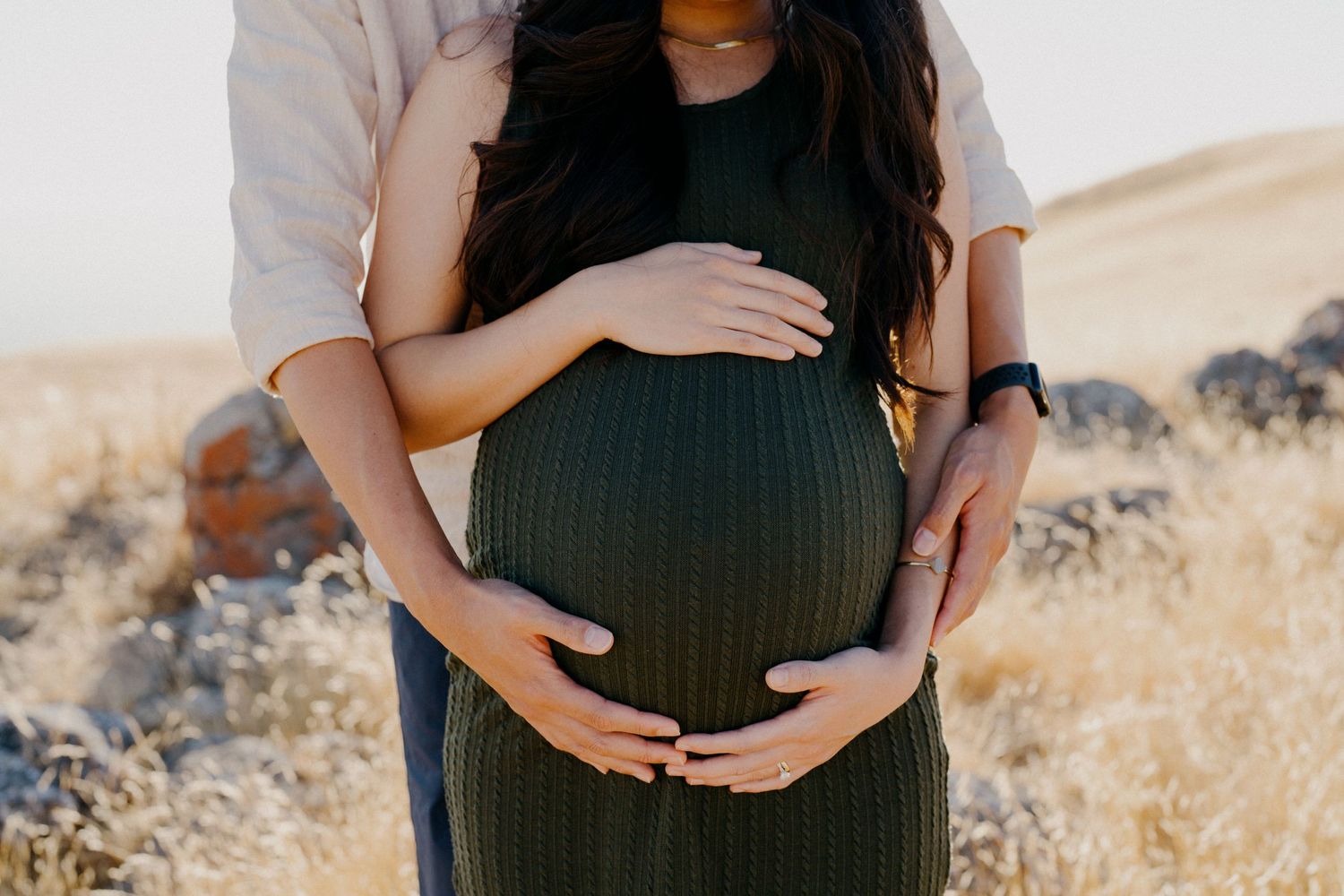 Close-up shot of hands cradling a pregnant belly in a sunlit field.