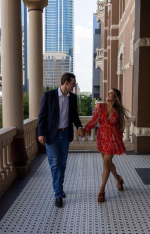 A couple walking hand in hand through a columned breezeway with downtown buildings visible in the background.