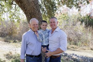 Three generations pose together under a large tree on a sunny day.