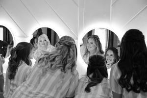 A black and white photograph of a bridal party getting ready together in a brightly lit room with mirrors.