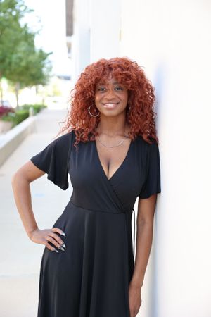 Stylish woman in black wrap dress with vibrant red curly hair poses against white wall.