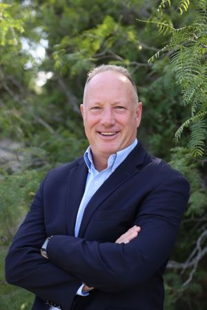 Professional headshot of person wearing navy blazer and light blue shirt against green foliage background.