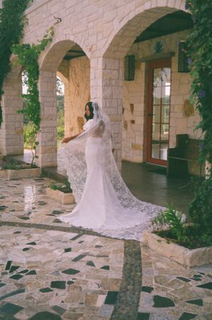 Elegant bride in a flowing lace veil poses under stone arches on a textured stone patio.