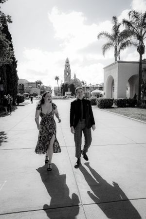 Black and white photo of couple walking along pathway with palm trees and Spanish architecture in background.
