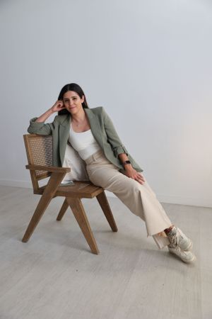 Woman in business casual attire lounges on wooden chair in minimalist white photography studio, The Lumen Room.