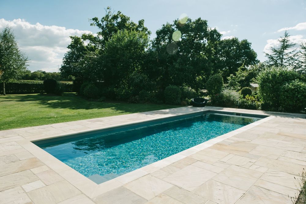 Rectangular swimming pool with stone patio surrounded by manicured lawn and trees.