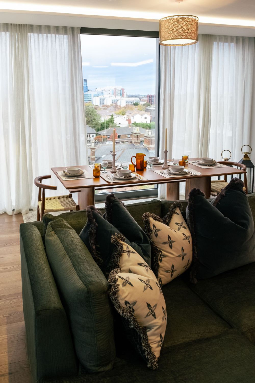 Modern dining area with green velvet sofa and city view through floor-to-ceiling windows.