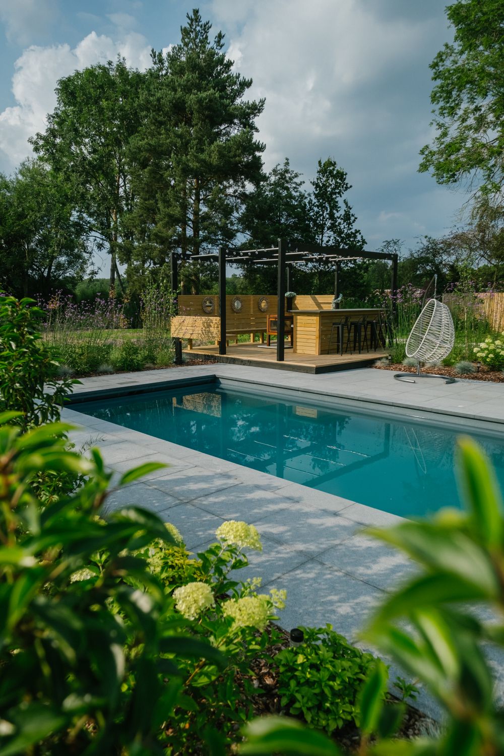 Modern pool area with pergola and hanging chair surrounded by lush garden landscaping.