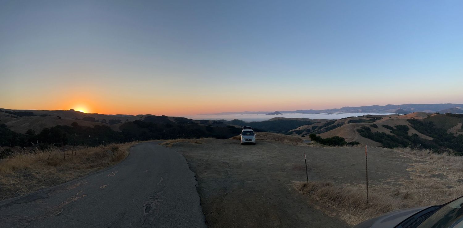 the sun sets behind a mountain while a layer of fog sits in the valley below