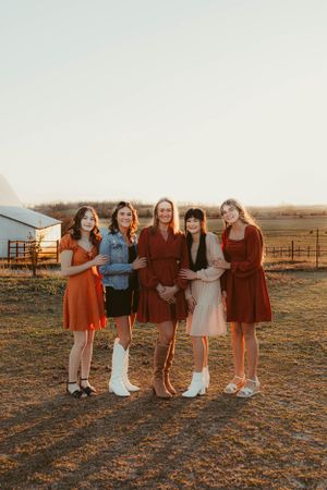 Group in vintage-inspired dresses poses together at sunset near a rustic barn in rural setting.