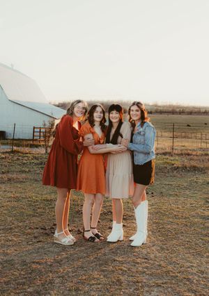Friends in autumn-colored dresses share a laugh during outdoor photoshoot by a blue barn.