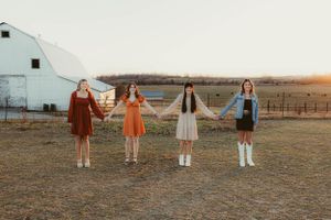 Four people holding hands in vintage-style dresses stand in line against rustic farm backdrop.