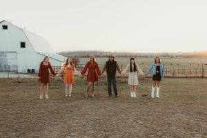 Group lineup holding hands against sunset farmland backdrop with white barn in background.