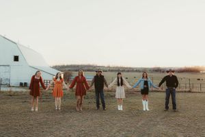 Line of people holding hands in vintage outfits pose in front of rustic barn at golden hour.