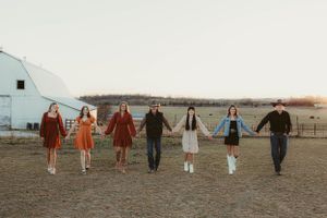 Group holding hands in retro attire poses in row against rural farm setting at sunset.