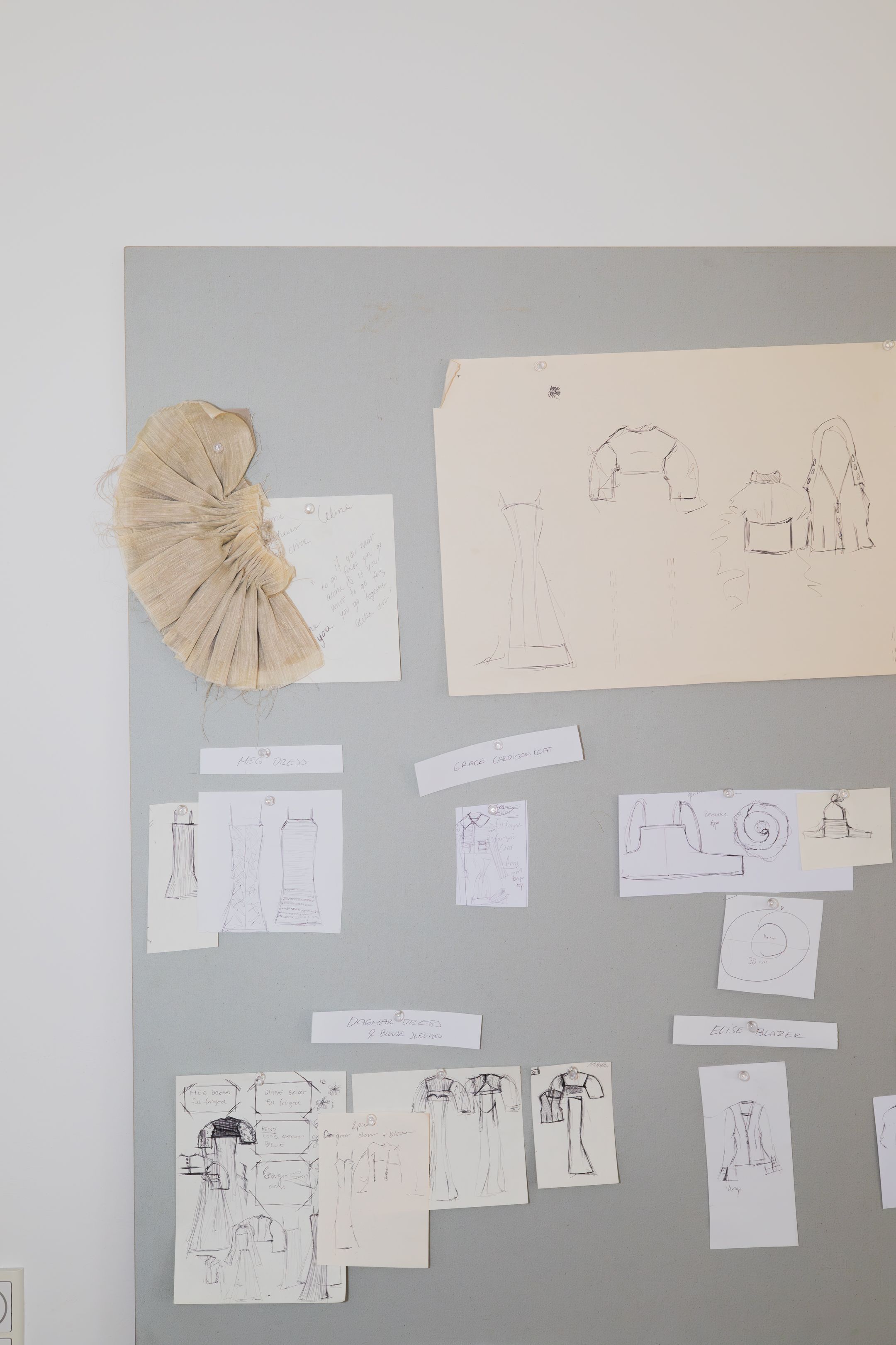 Collection of sketches and design drawings pinned to grey wall with fabric sample.