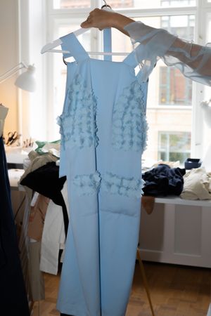 Light blue sleeveless dress with textured floral applique details hanging on wooden hanger by window.