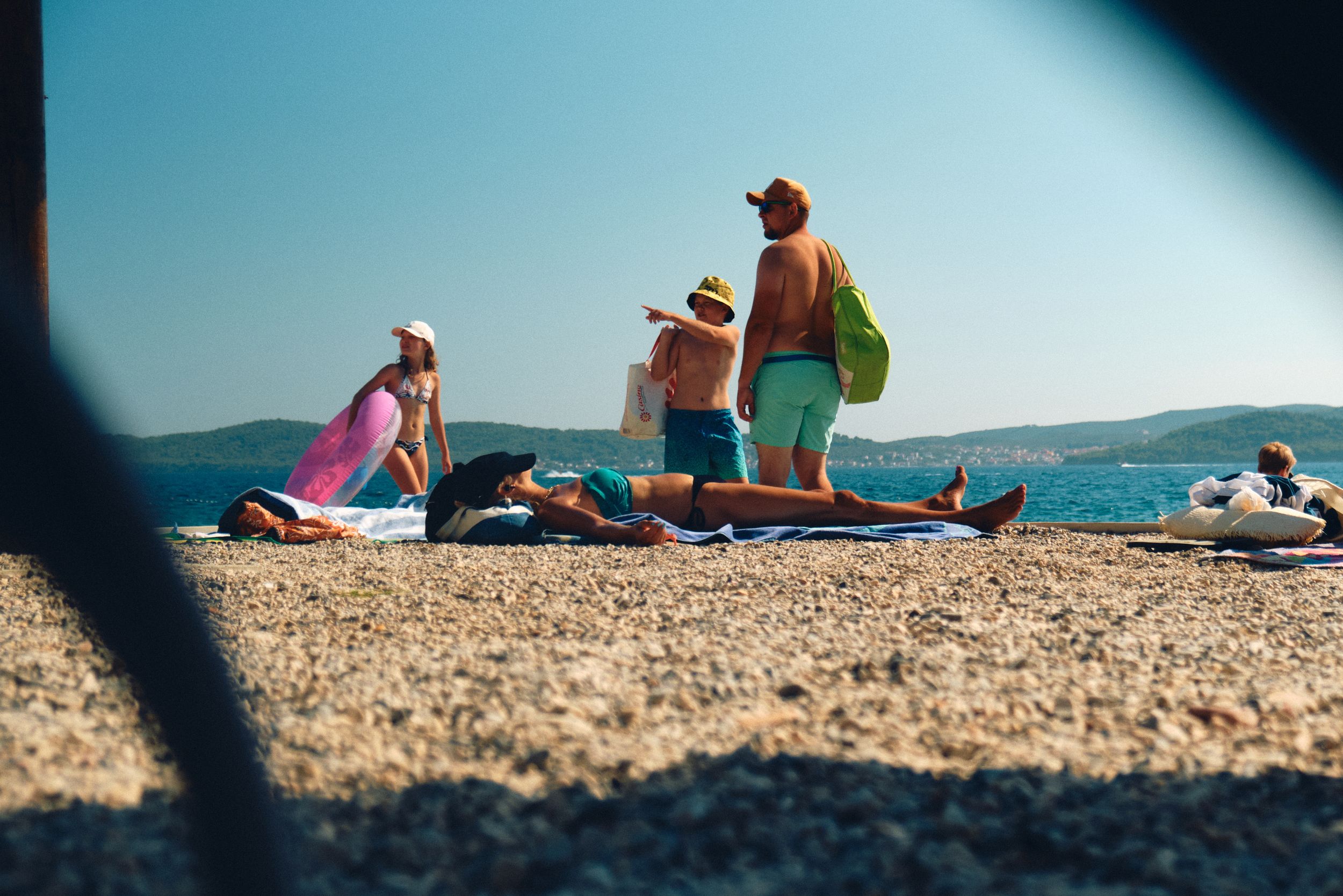 Beachgoers relaxing on a rocky shoreline with blue waters and mountains in the distance.