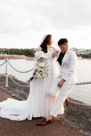 A couple in elegant white attire poses for wedding photos along a waterfront boardwalk at sunset.