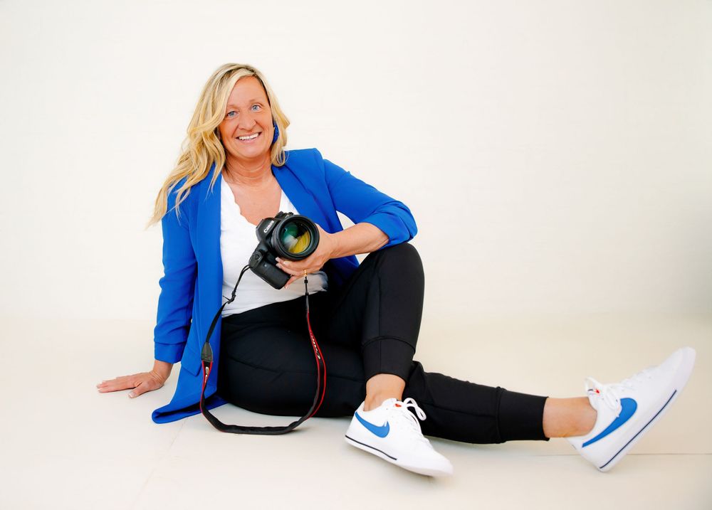 Photographer in bright blue blazer and black pants sits on the floor holding her camera against white background.