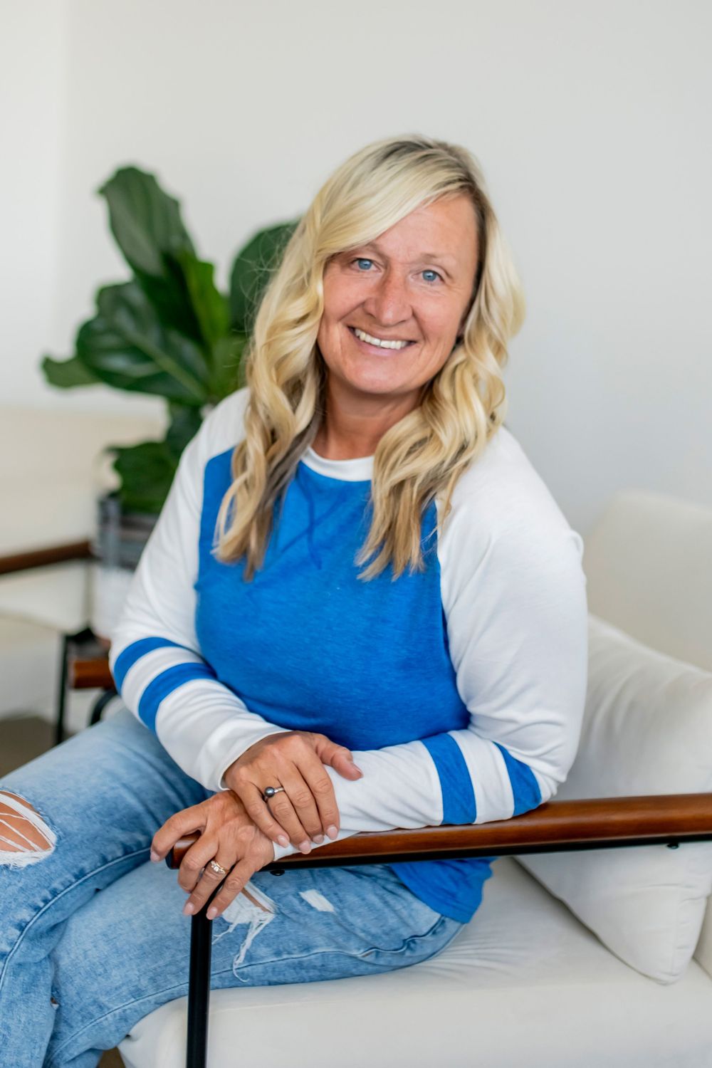 Casual portrait of business owner wearing a blue and white striped shirt sitting on a white chair with a plant in background.