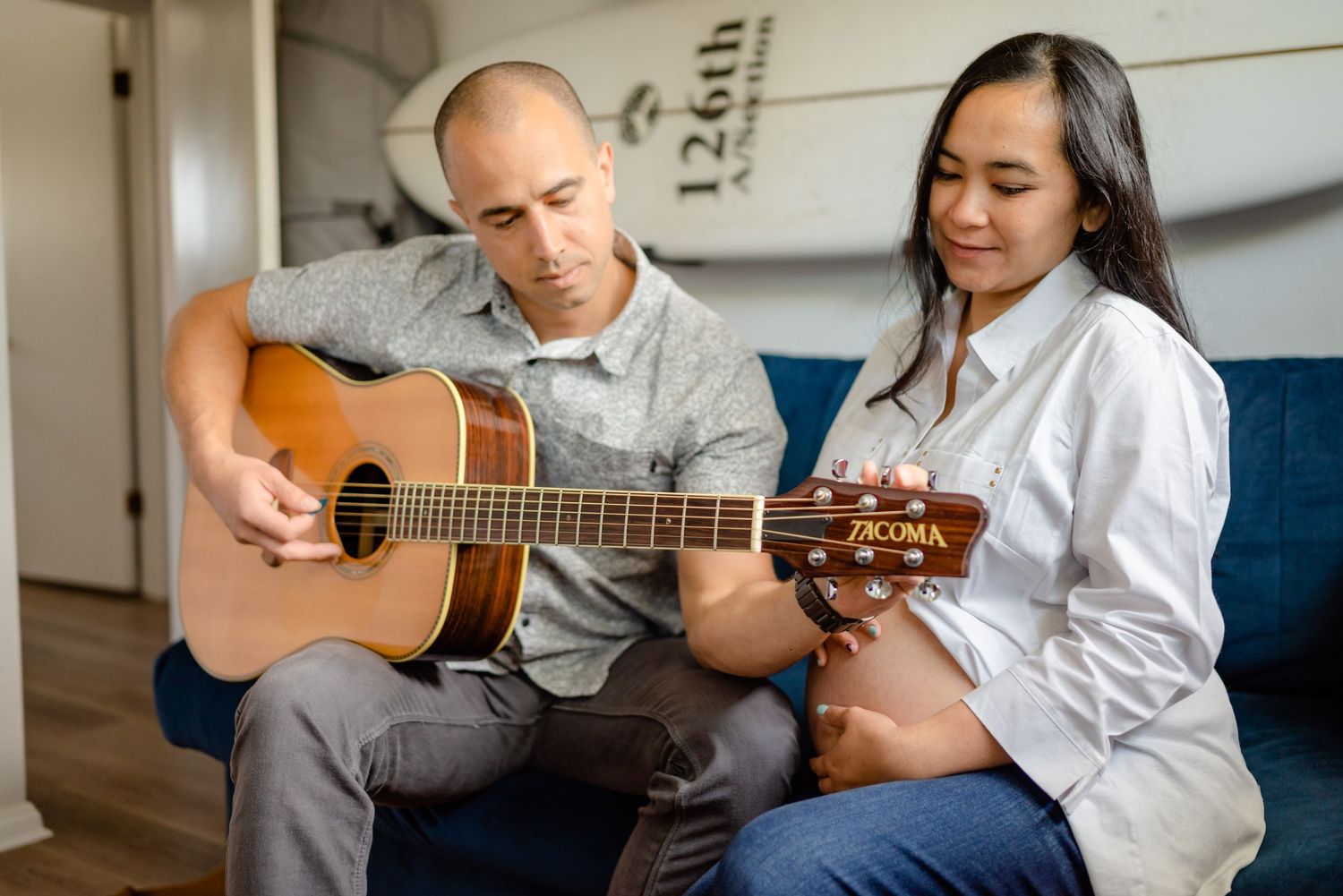 Man sitting on a blue couch with surf board hung on wall, plays guitar to his pregnant wife. In-home maternity session. Doughman Photography