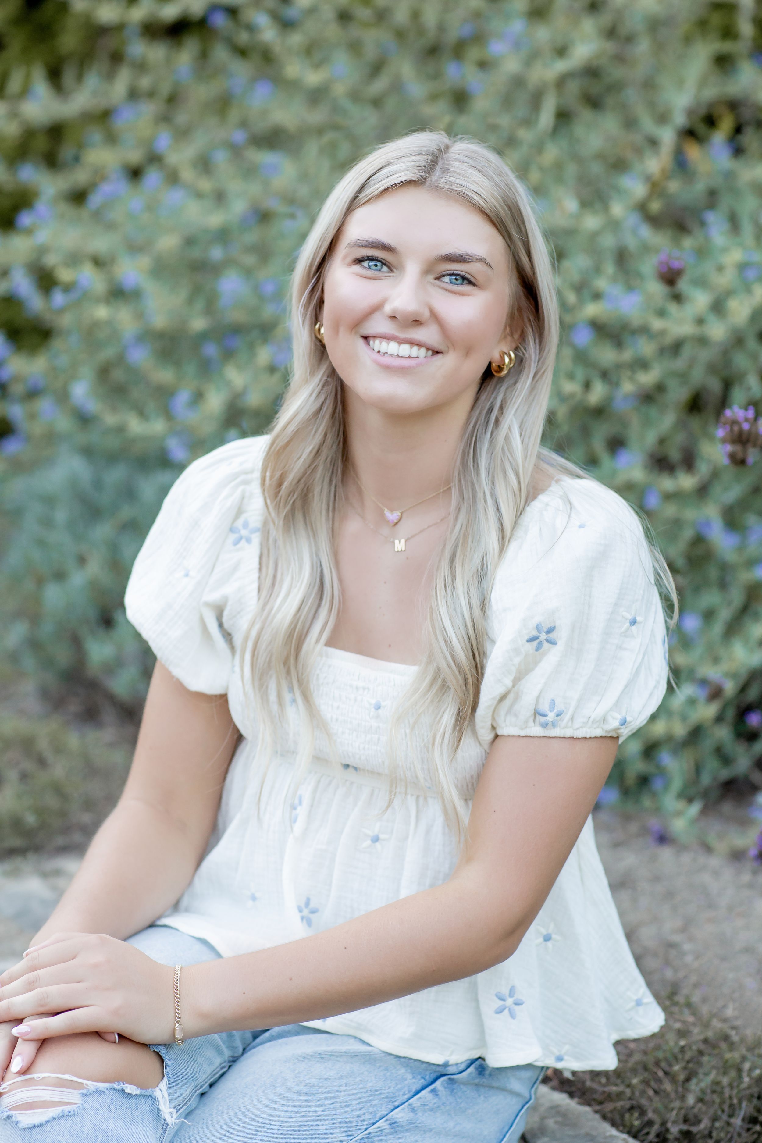 Smiling blonde woman in white floral top seated outdoors with blue flowering shrubs behind her.