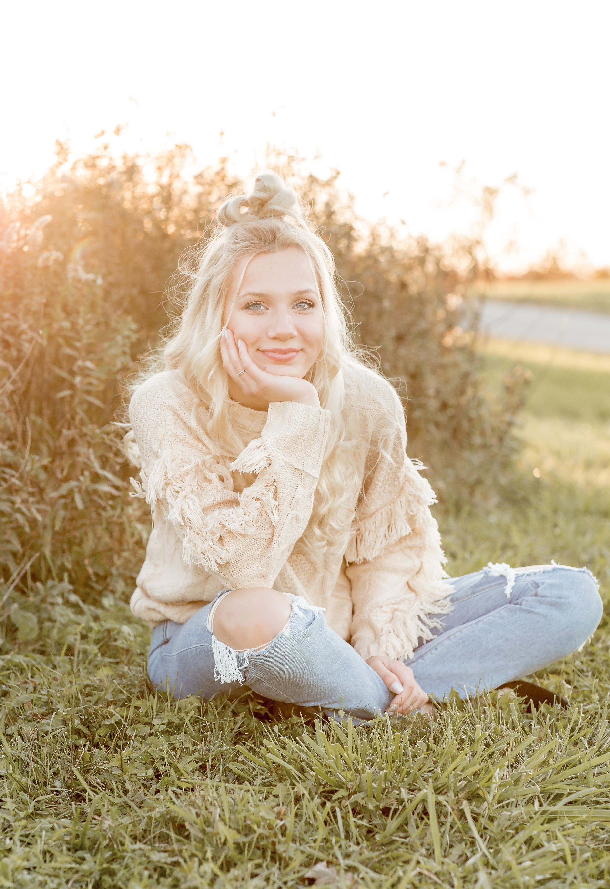 Blonde teenage girl sits cross-legged in golden field grass wearing cream fringe jacket at sunset.