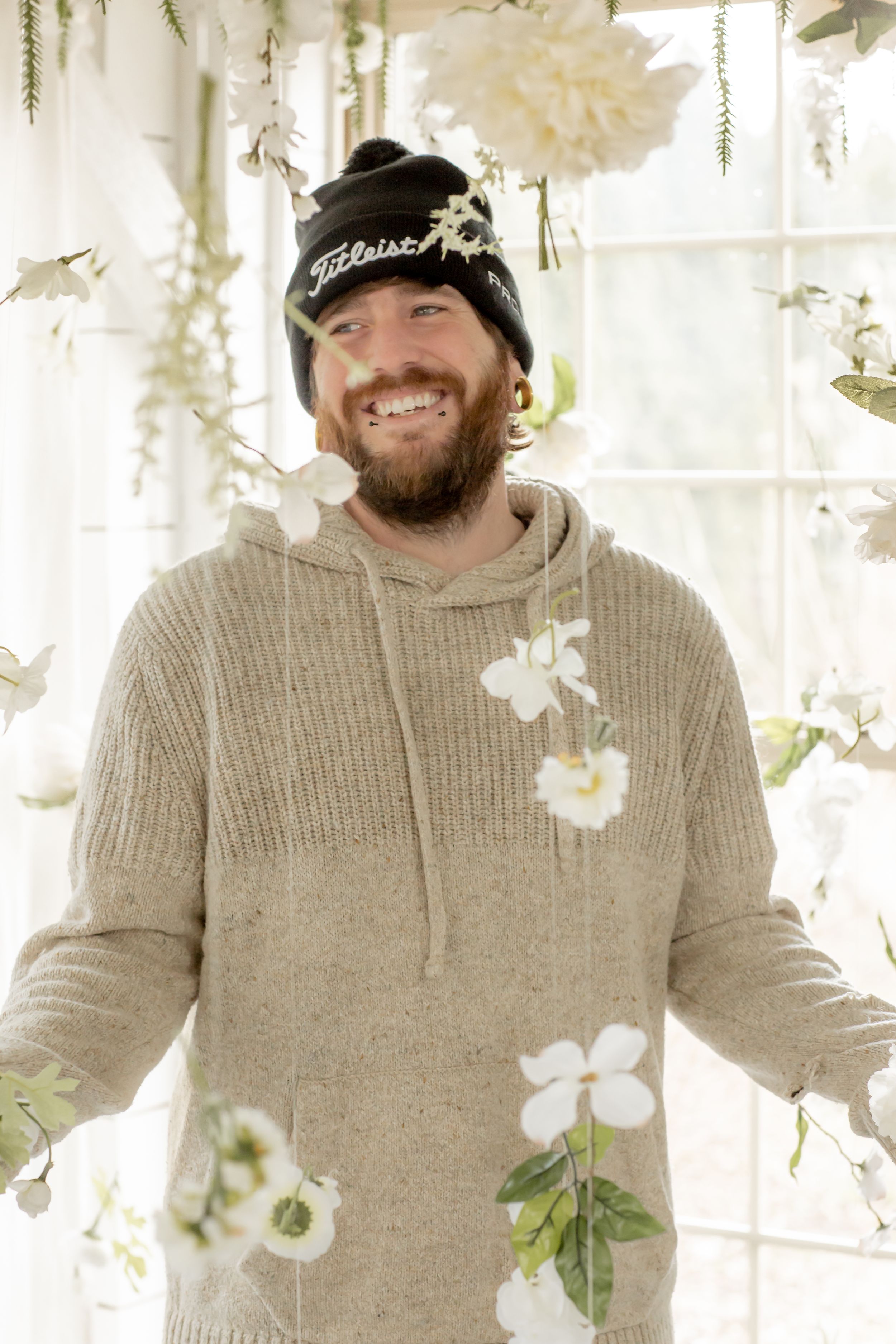 Bearded man in beige hoodie and Titleist beanie smiling among white hanging flowers.