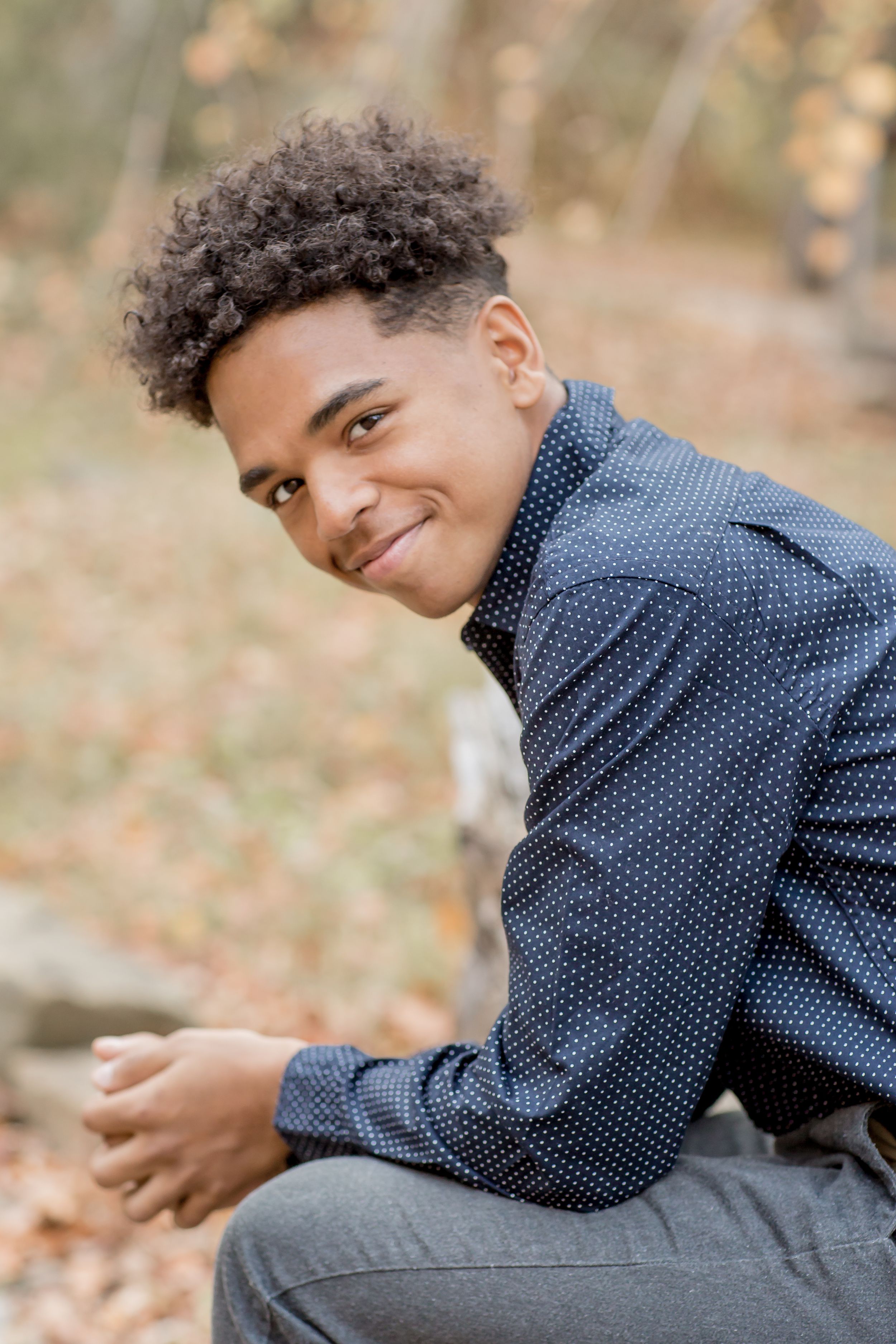 Smiling teenage boy in navy dotted shirt sitting on rocks among fall foliage.