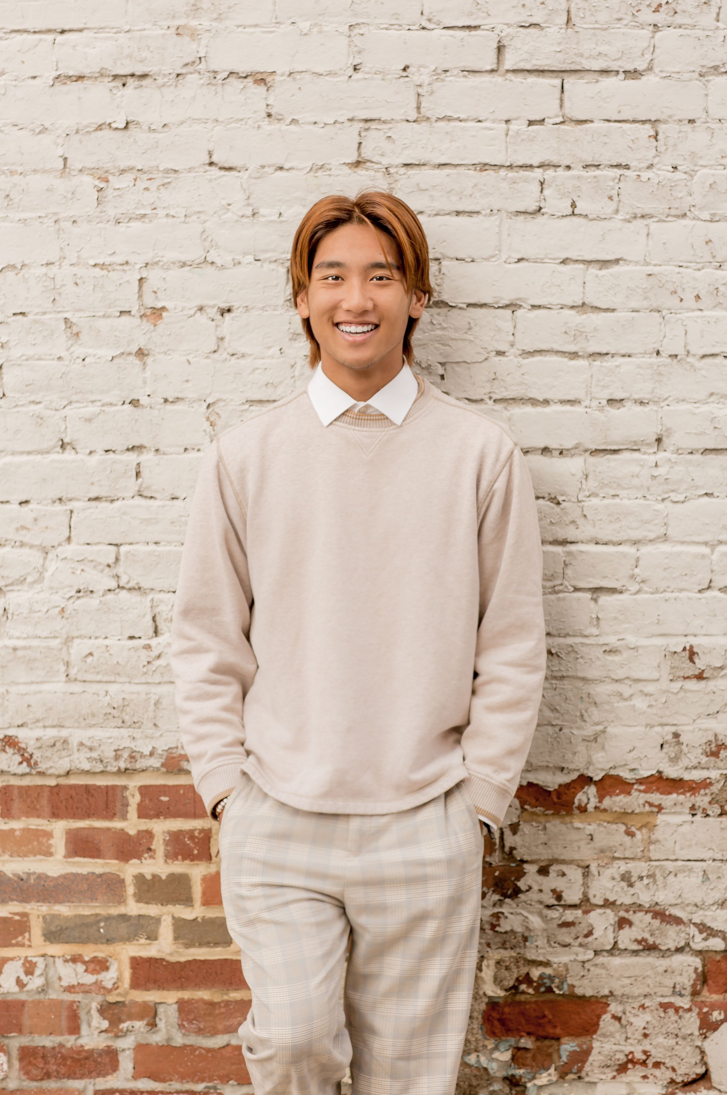Smiling young man in beige sweater standing against white painted brick wall outdoors.