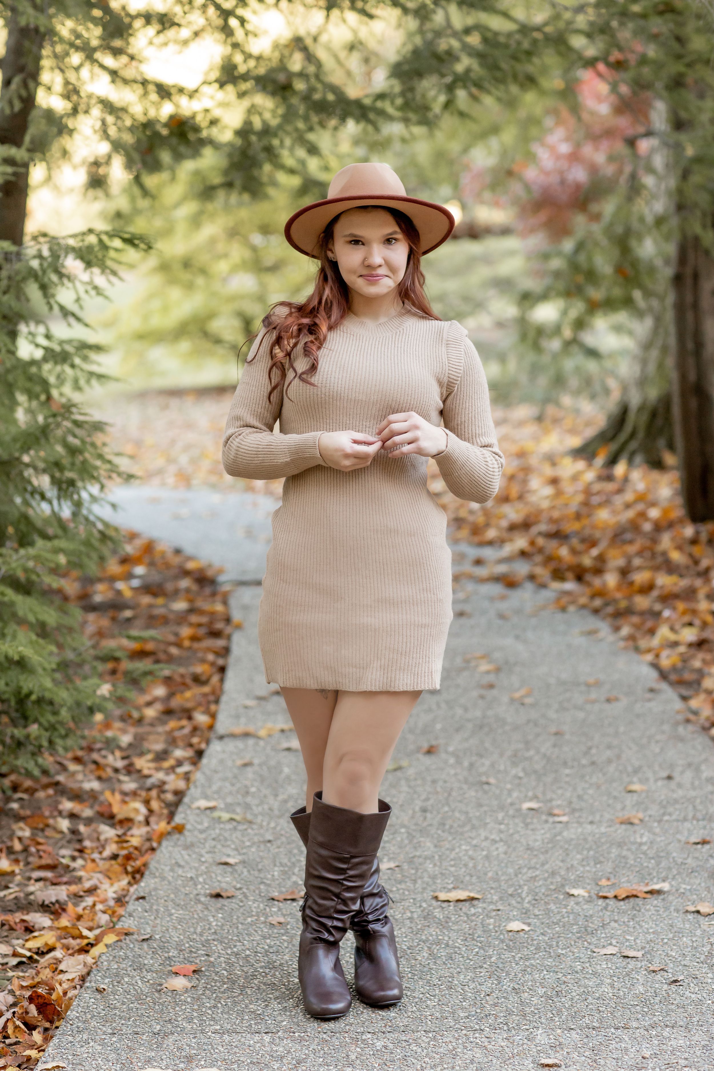 Young woman in tan sweater dress and brown boots wearing wide-brim hat on autumn pathway.