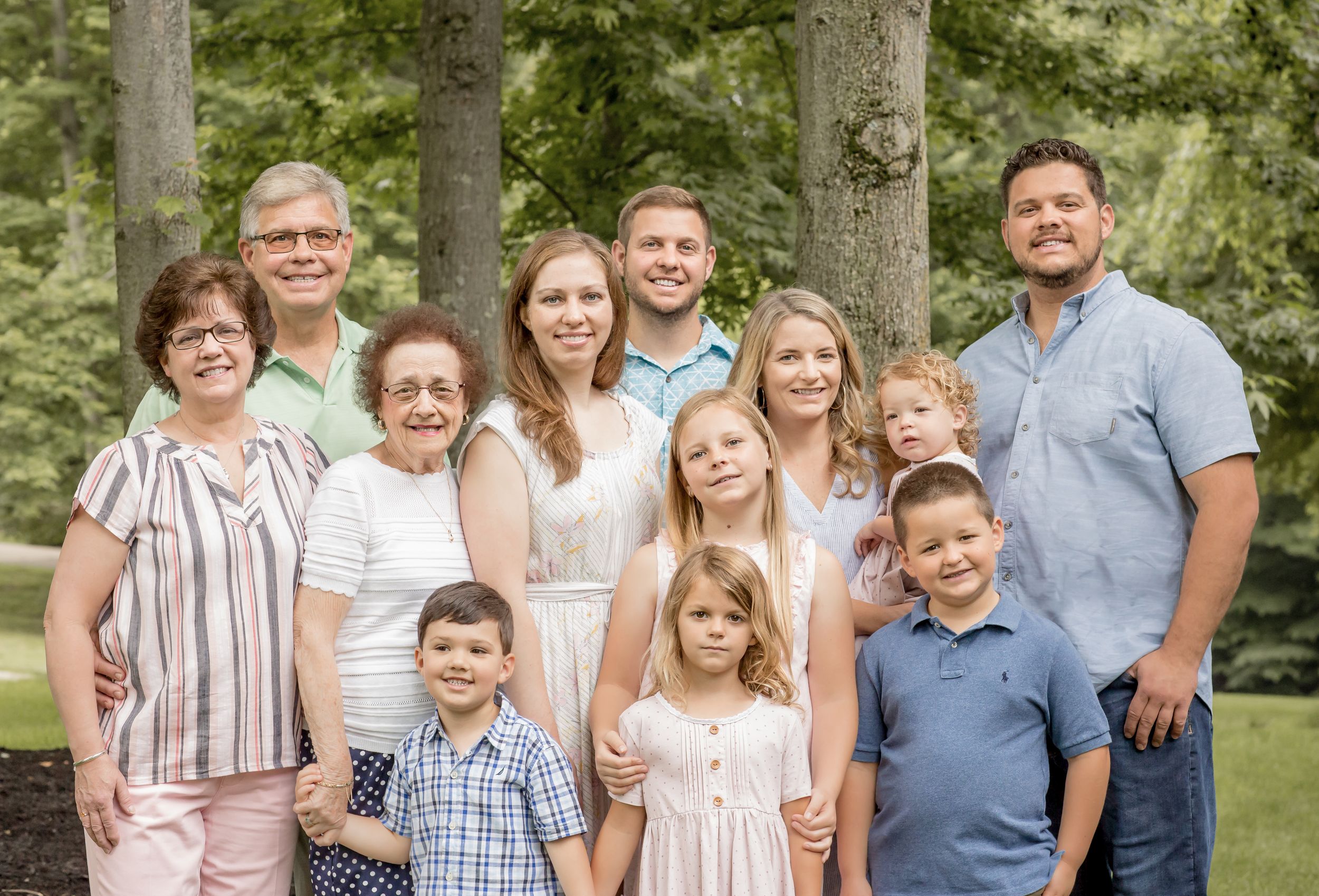 Large multigenerational family smiling together outdoors surrounded by tall green trees.