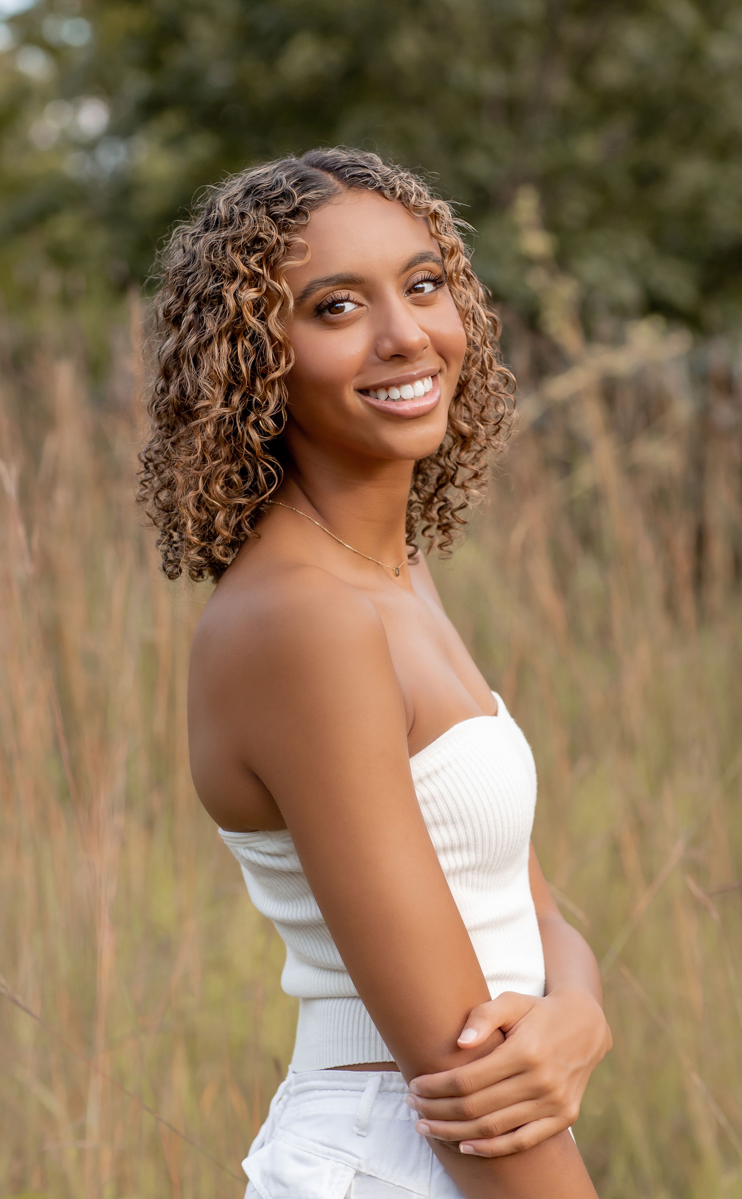 Young woman with curly hair in white tube top smiling in a golden grass field.