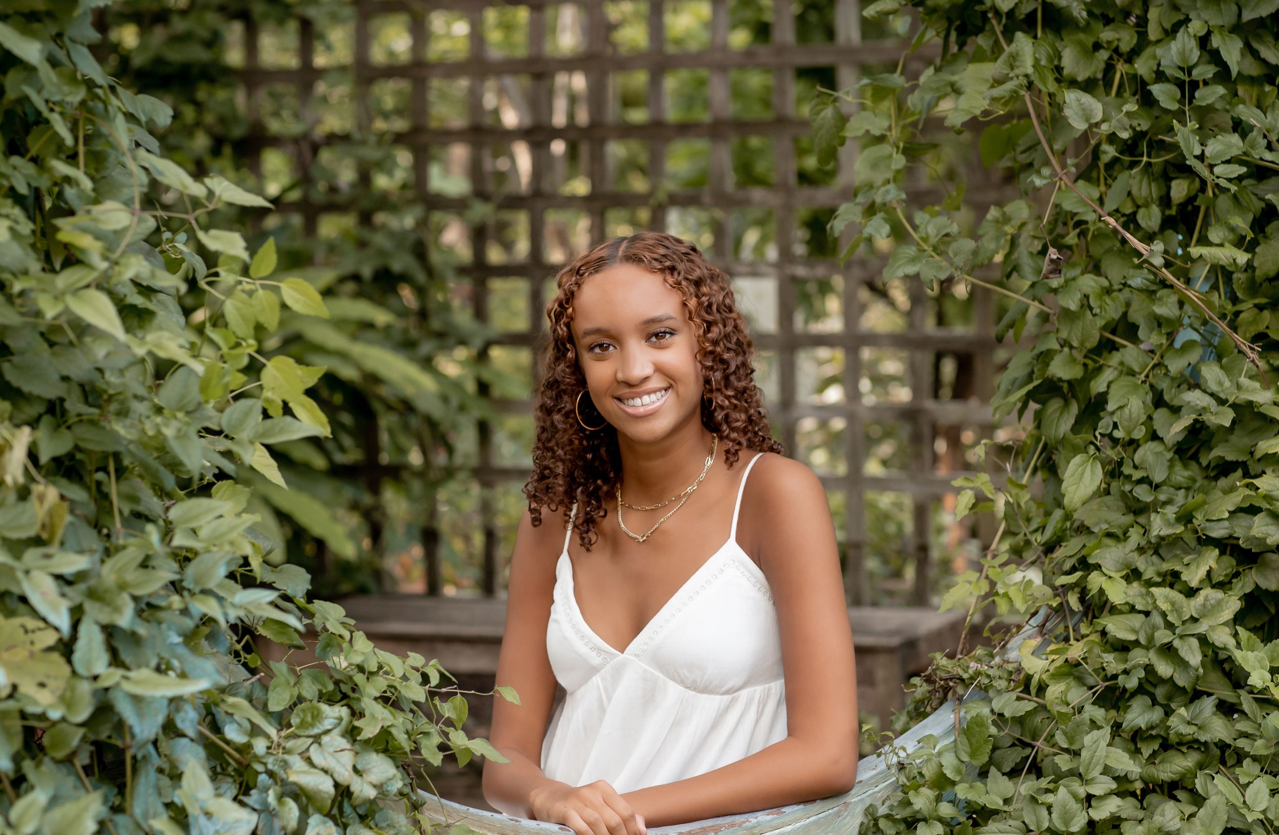 Young woman in white dress smiling in front of a vine-covered wooden trellis.