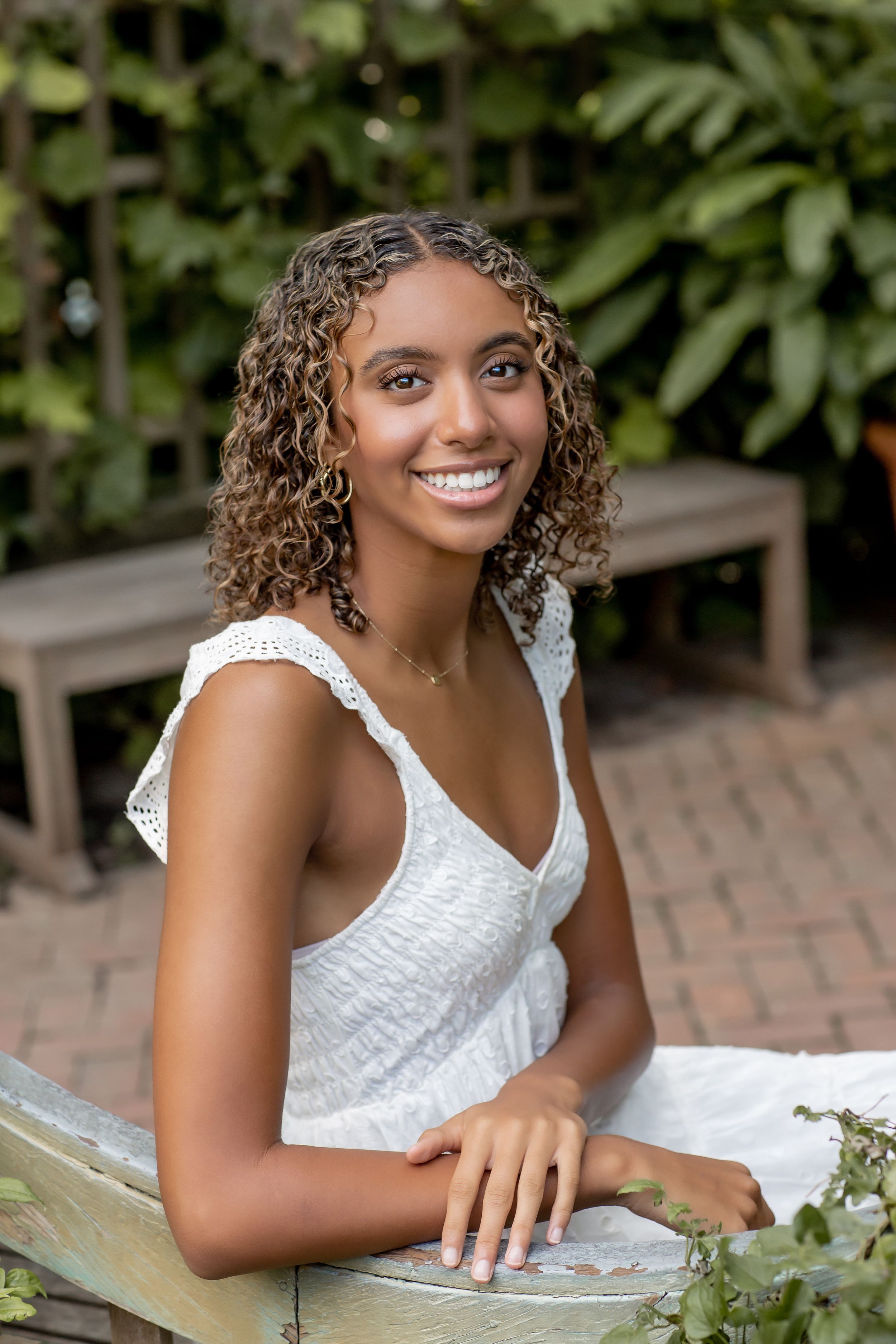 Young woman with curly hair in white sundress smiling at camera in lush garden setting.