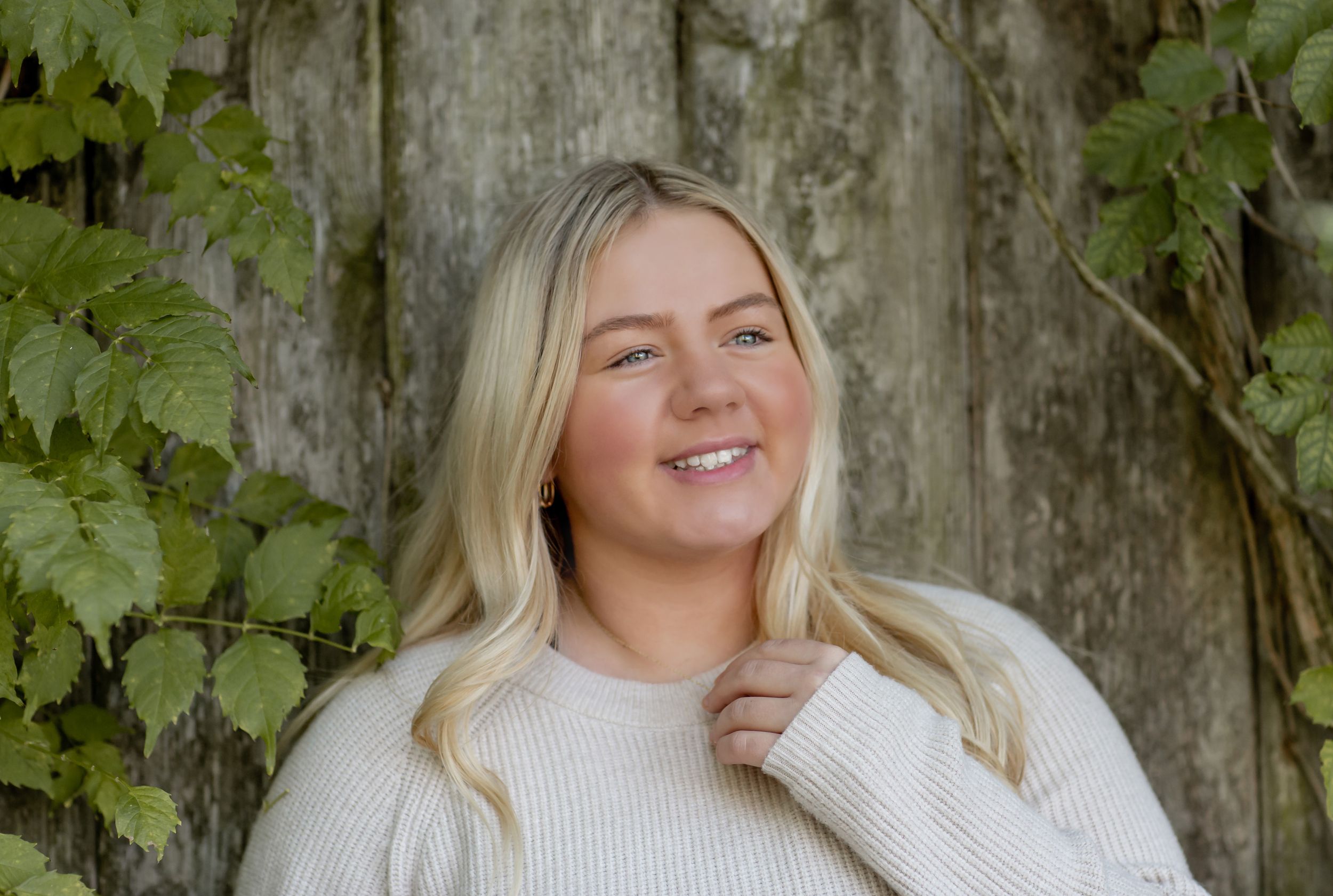 Smiling blonde woman in cream sweater leaning against rustic wooden wall with ivy.