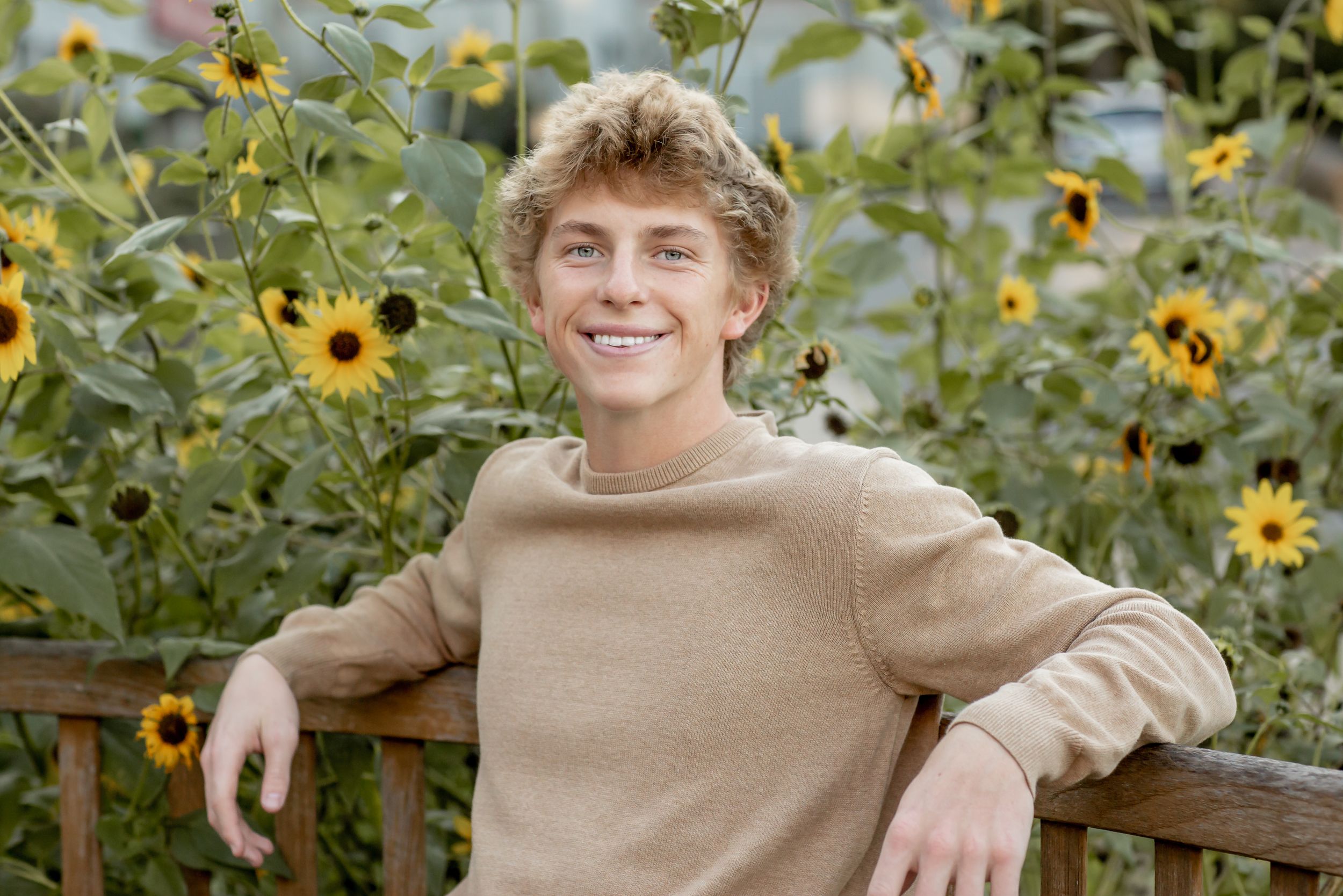 Smiling teenage boy in beige sweater leaning on a fence with sunflowers behind him.