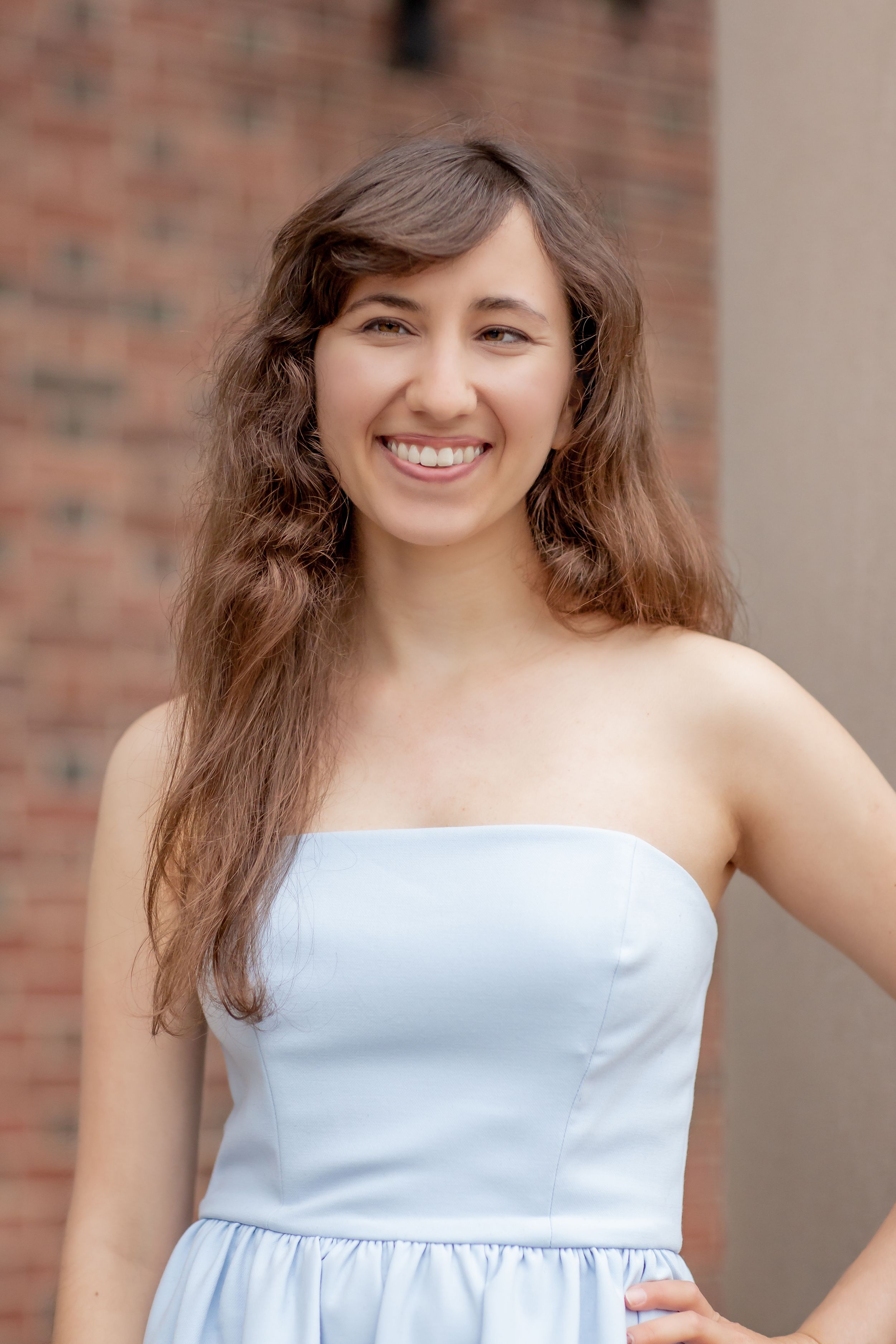 Smiling young woman in light blue strapless dress posing in front of brick wall.