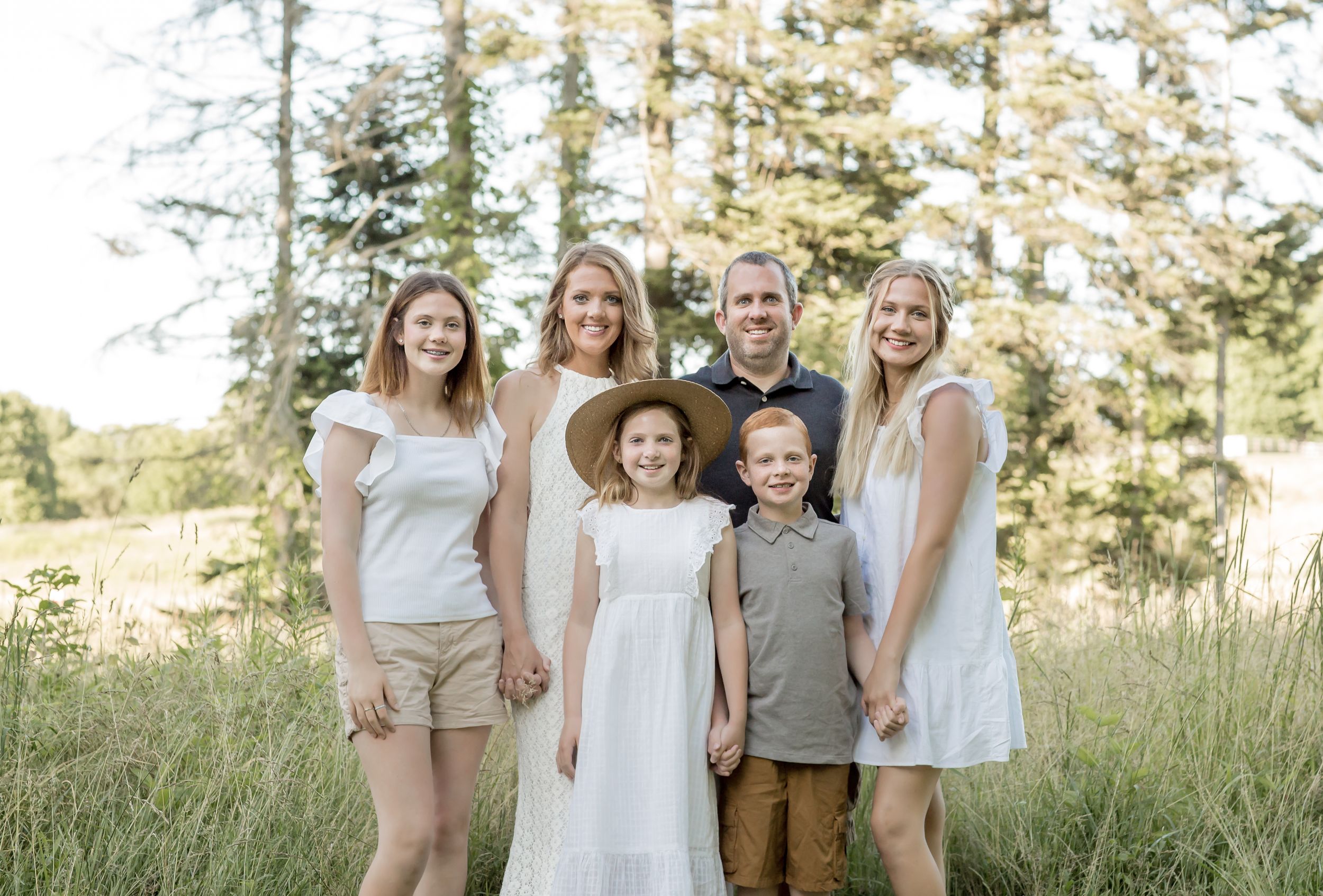 Happy family of six posing in a meadow surrounded by tall pine trees in warm light.